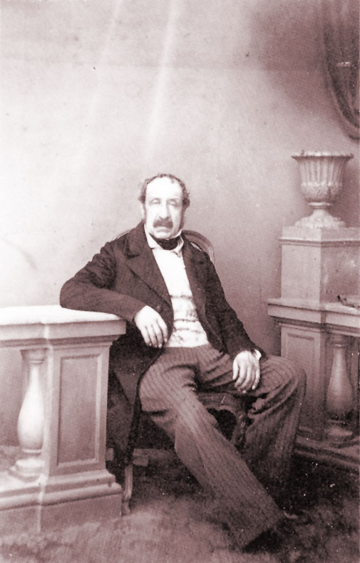 Ritratto fotografico ottocentesco in tonalità seppia di Filippo Bruti Liberati, in abito formale scuro con panciotto, accanto a un piedistallo con anfora decorativa.