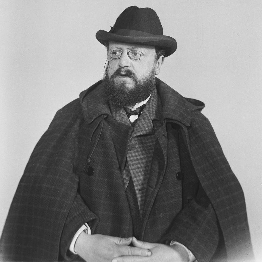 Ritratto fotografico in bianco e nero di Alessandro Bruti Liberati, di stile Belle Époque, con barba folta, occhiali tondi con catenella, cappello di feltro e mantellina di panno a quadri scozzesi.
